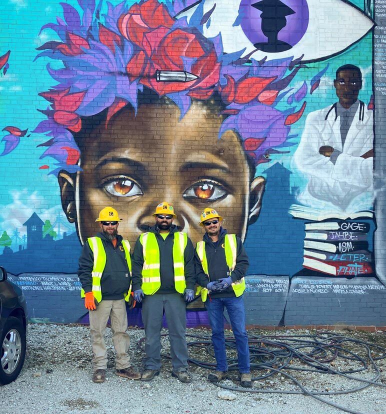 Three Commonwealth employees stand in front of the mural at Creative Visions in Des Moines, IA.